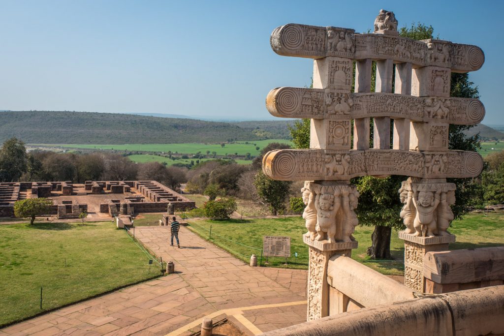 The western gateway (torana) of Stupa 1 at the Great Stupa of Sachi is supported by pot-bellied figures. In the distance is the foundation of Monastery 51. Sanchi is set in an idyllic rural setting and it provided a respite from the busy cities. (© Richard McGuire photo)